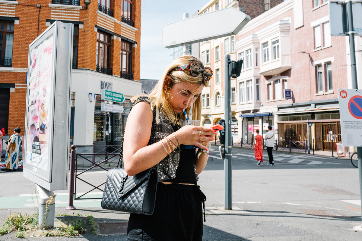 Une jeune femme avec son smartphone et un sac noir Yves Saint Laurent boit une boisson avec une paille à Lille