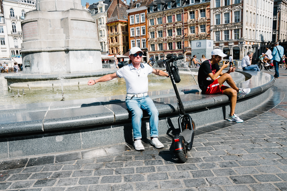 Un homme assis sur la fontaine de la Déesse sur la Grand place de Lille étend ses bras à côté de sa trotinette.