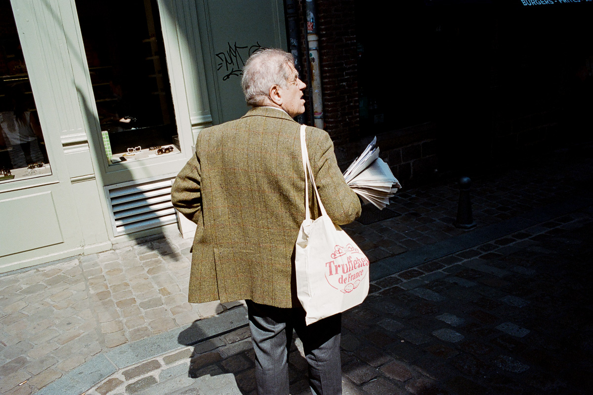 Un homme avec un tote bag et les bras chargés de journaux cherche sa route place des Patiniers à Lille