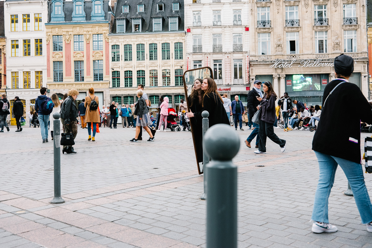 Une jeune femme porte un miroir et traverse la Grand place de Lille.